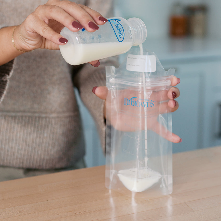 Person pouring formula from a Dr. Brown's bottle into a clear pouch on a wooden surface.