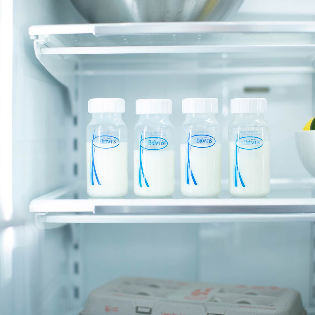 Four Dr. Brown's™ Breast Milk Collection Bottles from Dr. Brown's, filled with milk, are lined up on a refrigerator shelf beside a white bowl holding a lemon, with a carton of eggs below.