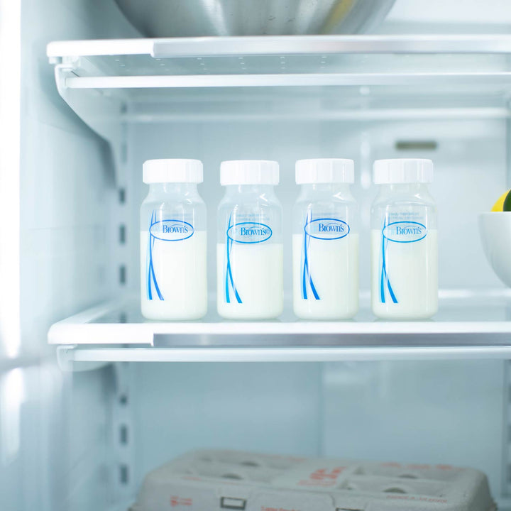 Four Dr. Brown's™ Breast Milk Collection Bottles from Dr. Brown's, filled with milk, are lined up on a refrigerator shelf beside a white bowl holding a lemon, with a carton of eggs below.
