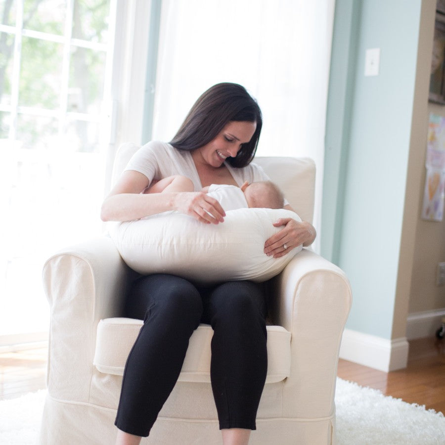 A woman sits in a comfortable cream-colored chair in a softly lit room, breastfeeding a baby using a supportive nursing pillow. Sunlight filters through a nearby window, creating a warm and serene atmosphere.