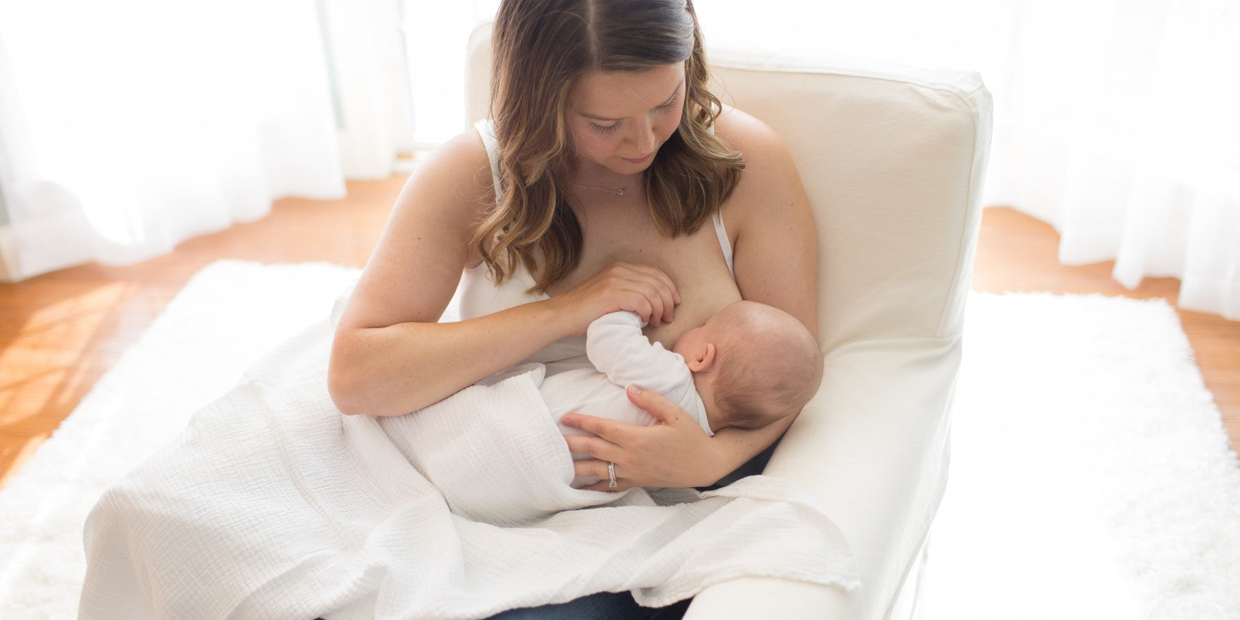 A woman is sitting on a white chair breastfeeding a baby wrapped in a white blanket. The room has soft lighting with white curtains and a light carpeted floor.