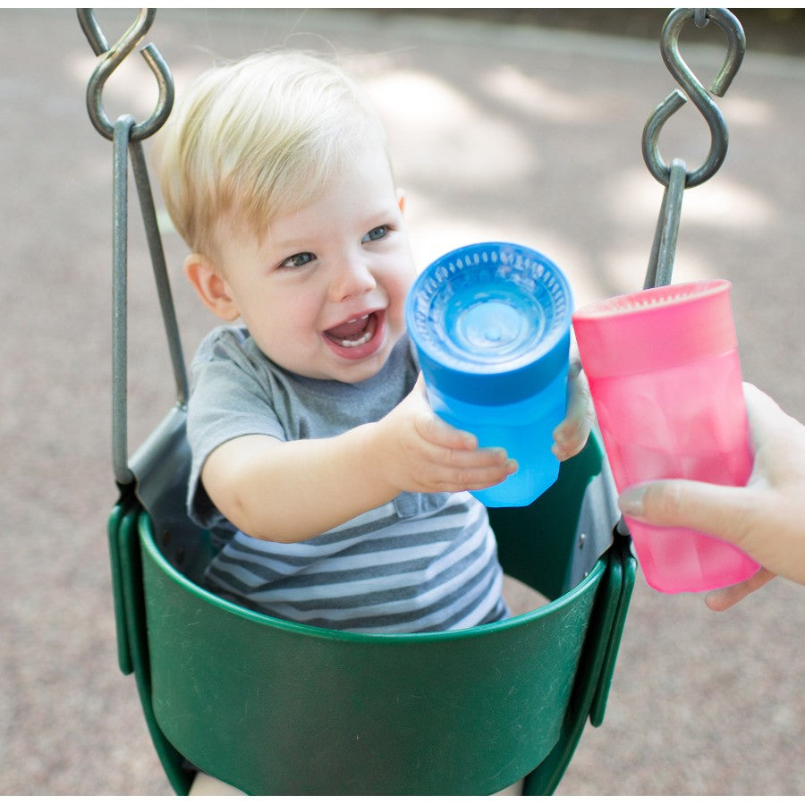 A smiling toddler with blonde hair sits in a green swing, reaching for a blue sippy cup from an adult's hand holding a pink cup. The background is a blurred outdoor playground setting.