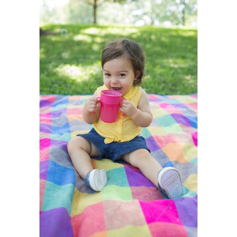 A toddler wearing a yellow top and denim shorts sits on a colorful plaid blanket in a grassy area, holding and drinking from a pink cup. The background has greenery and blurred trees.