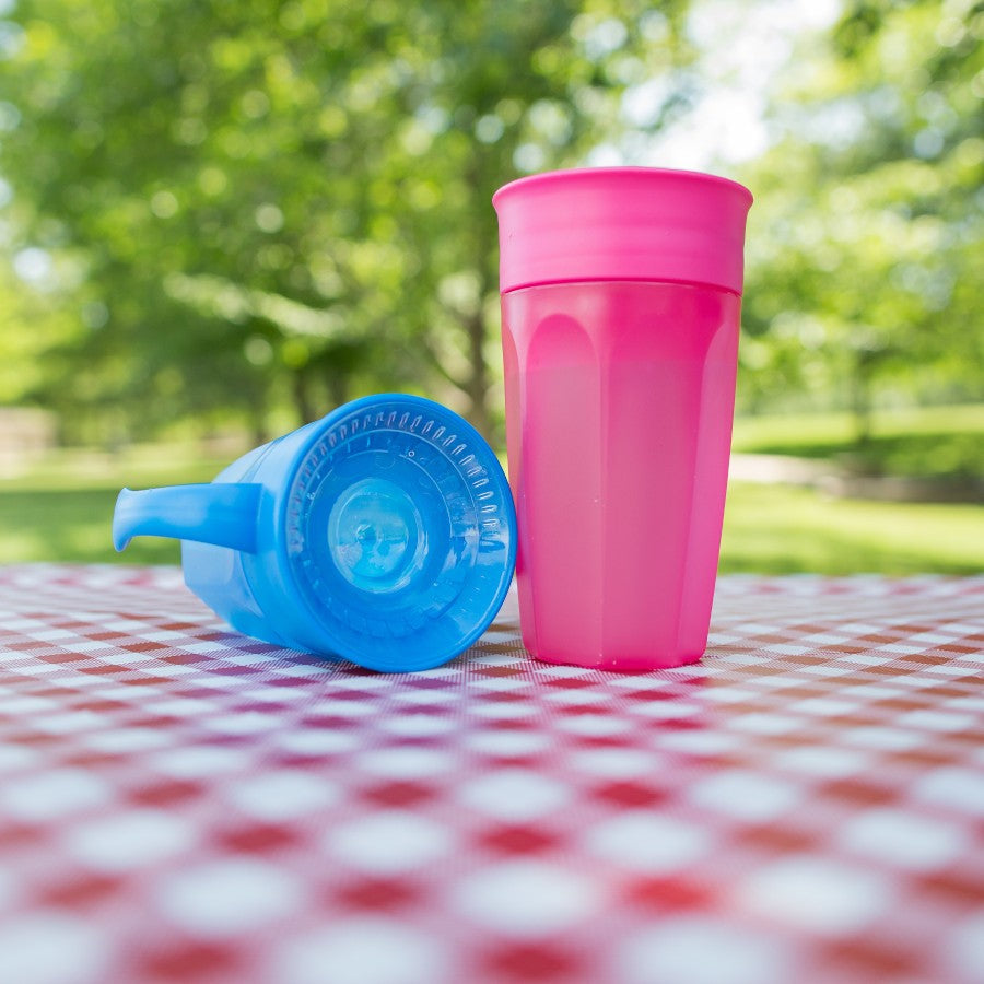 Two colorful plastic sippy cups, one pink standing upright and one blue on its side, are placed on a red and white checkered tablecloth. The background features a sunny park with green trees and grass.