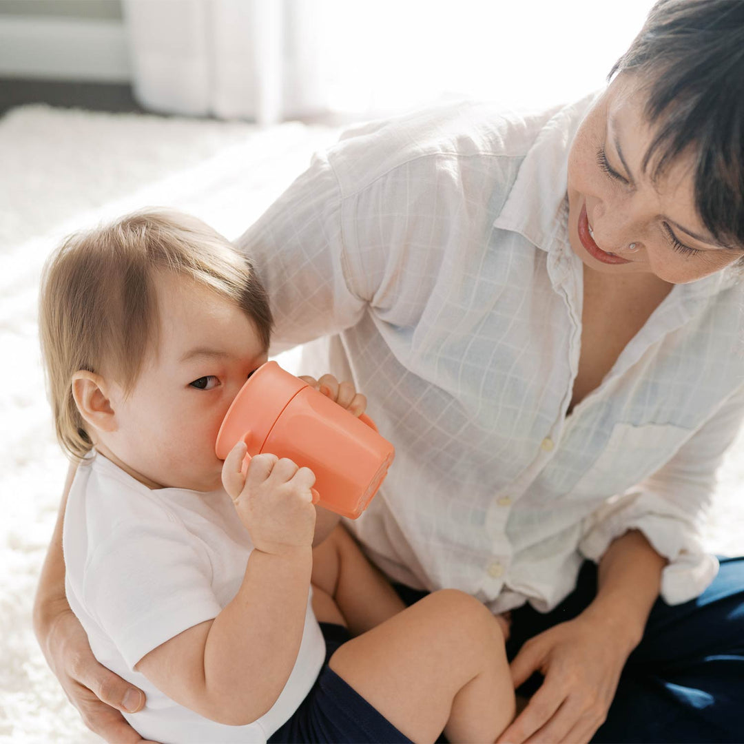 Woman holding a child who is drinking from a coral-colored Cheers360 cup indoors.