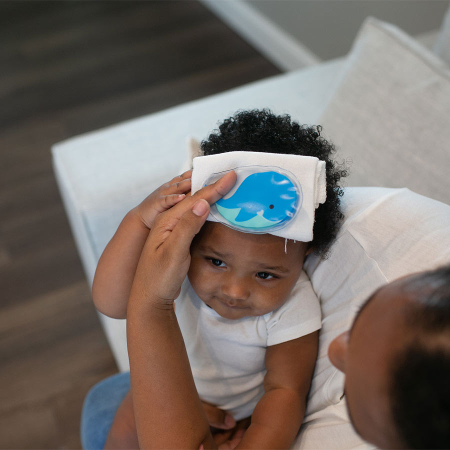 A caregiver applying a blue ice pack with a whale design to a baby's head in a living room. The baby, wearing a white shirt, is seated on the caregiver's lap, resting on a white sofa.