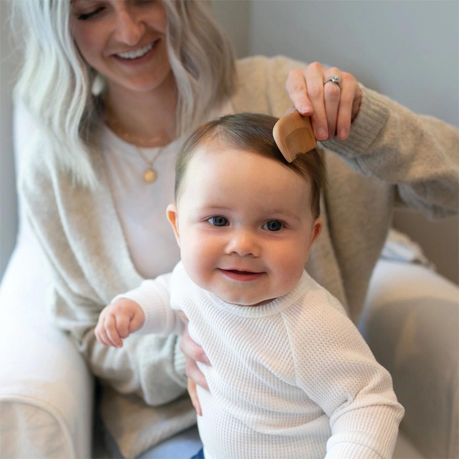 A smiling baby in a white outfit sits on a woman's lap, while she gently combs the baby's hair with a wooden brush. The woman, wearing a light sweater, is partially visible and smiling. The background is a softly lit room.