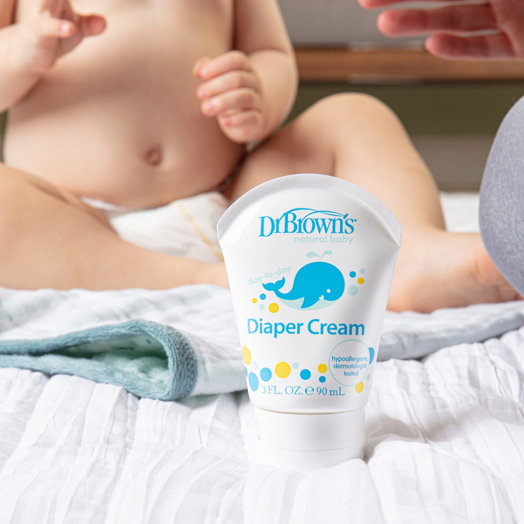 A baby sits on a changing mat, reaching towards a tube of Dr. Brown's Diaper Cream in the foreground. The cream is labeled as hypoallergenic and dermatologist tested. The baby is wearing a diaper, and a blanket is partially visible.