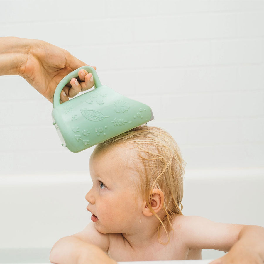 A baby in a bathtub looks up while water is gently poured over their head from a green pitcher with a handle and leaf designs. The background features white tiles.