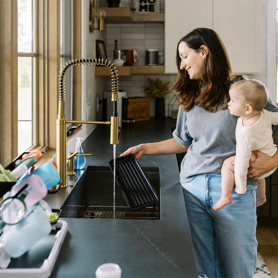 A woman holding a baby washes a tray under a faucet in a modern kitchen. The countertop is cluttered with dishes and a dish rack is visible. Natural light streams through a large window.