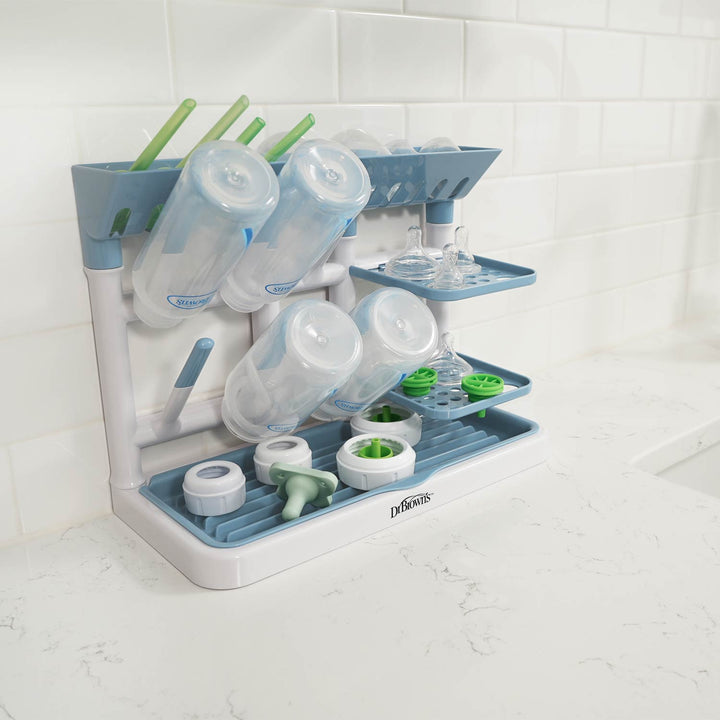 A light blue and white baby bottle drying tower on a white marble countertop against a white subway tile backsplash. The rack is shown holding several baby bottles, cups, and straws.