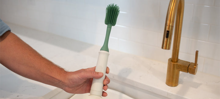 Hand holding a white and green cleaning brush near a sink with a gold faucet.