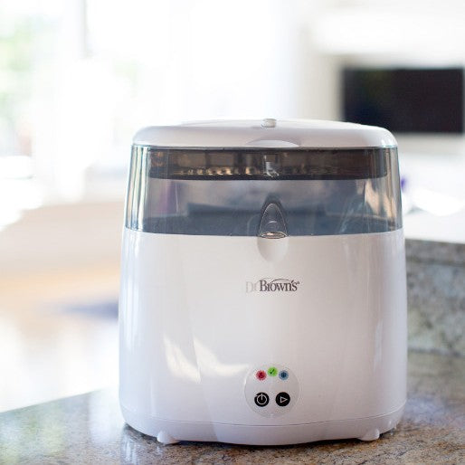 A white Dr. Brown's baby bottle sterilizer sits on a kitchen counter. The device has a transparent lid and simple control buttons on the front. Sunlight filters through a window in the background, creating a warm and bright atmosphere.