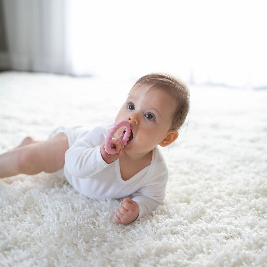 A baby wearing a white outfit lies on a soft, fluffy carpet. The baby is gazing upward and holding a pink teething toy in their mouth, with their left hand supporting their body. The background is softly lit, creating a cozy atmosphere.