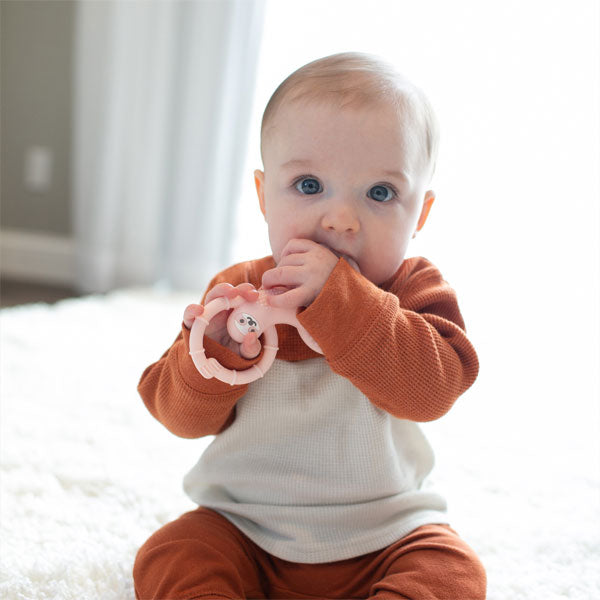 A baby with blue eyes is sitting on a soft white rug, wearing a brown and cream outfit. The baby is holding a pink teething ring and is putting it in their mouth. The background shows a softly lit room with curtains.