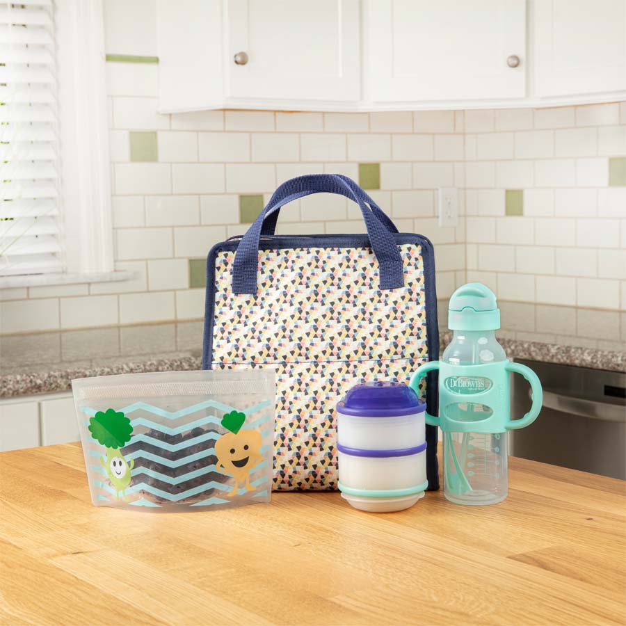 A kitchen countertop featuring a patterned lunch bag, a reusable snack bag with cute vegetable illustrations, a stackable container, and a turquoise water bottle with a handle. The background includes a tiled backsplash and a window.