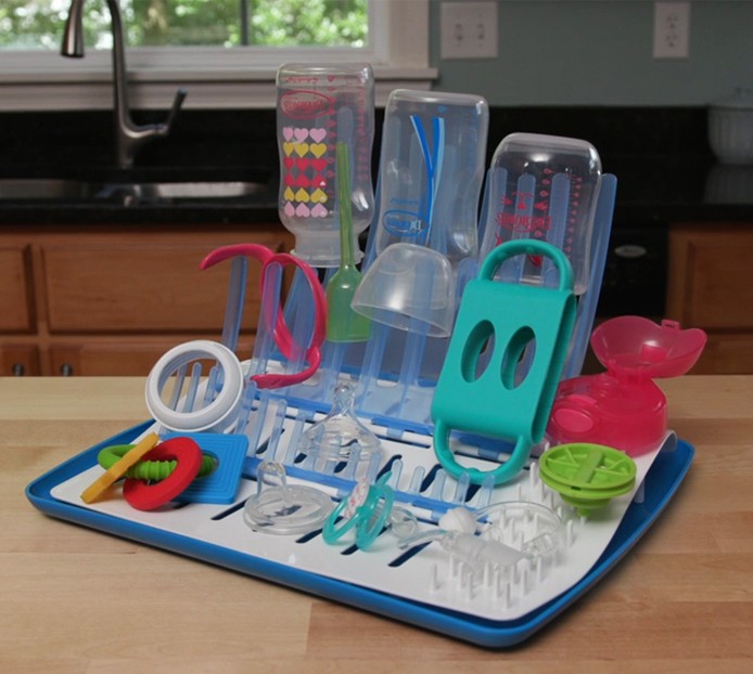 A countertop dish rack holds various colorful baby bottle parts, lids, and pacifiers. The kitchen setting includes a sink and wooden cabinets in the background. The dish rack is blue and white with several drying items upright.