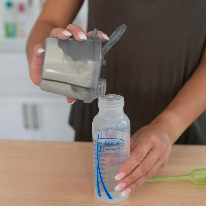 Person using a formula dispenser to pour formula into a bottle on a kitchen counter.