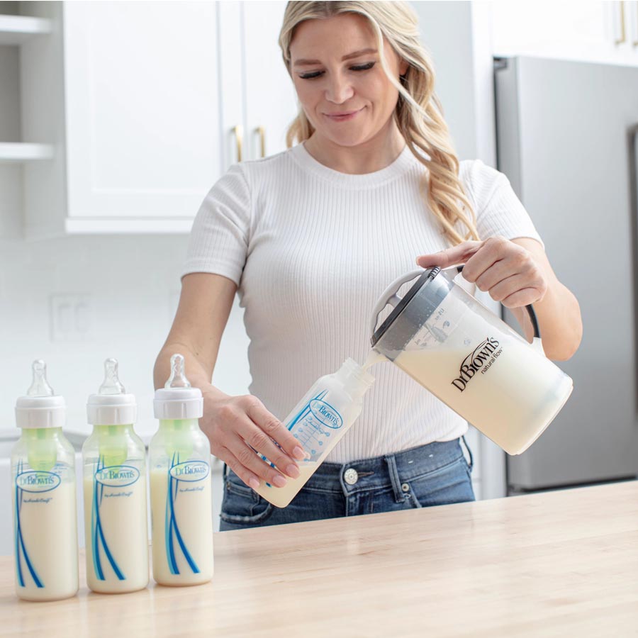 A person fills Dr. Brown's baby bottles with milk from a large container in a kitchen. The individual is wearing a white shirt and jeans, and is standing at a wooden counter with a few bottles in front of them.
