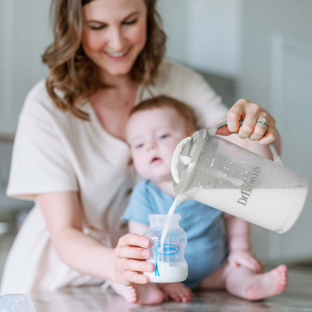 A smiling woman pours milk from the Dr. Brown’s® Formula Mixing Pitcher by Dr. Brown’s into a baby bottle while holding her baby seated on the kitchen counter.