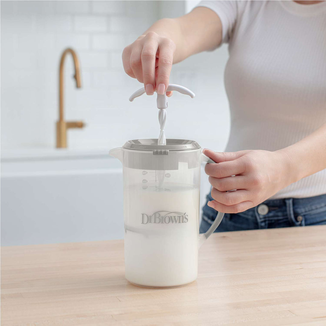Wearing a white shirt, someone prepares formula with Dr. Brown's® Formula Mixing Pitcher by Dr. Brown's on a light wood counter, baby bottles at hand and a gold faucet in the background.