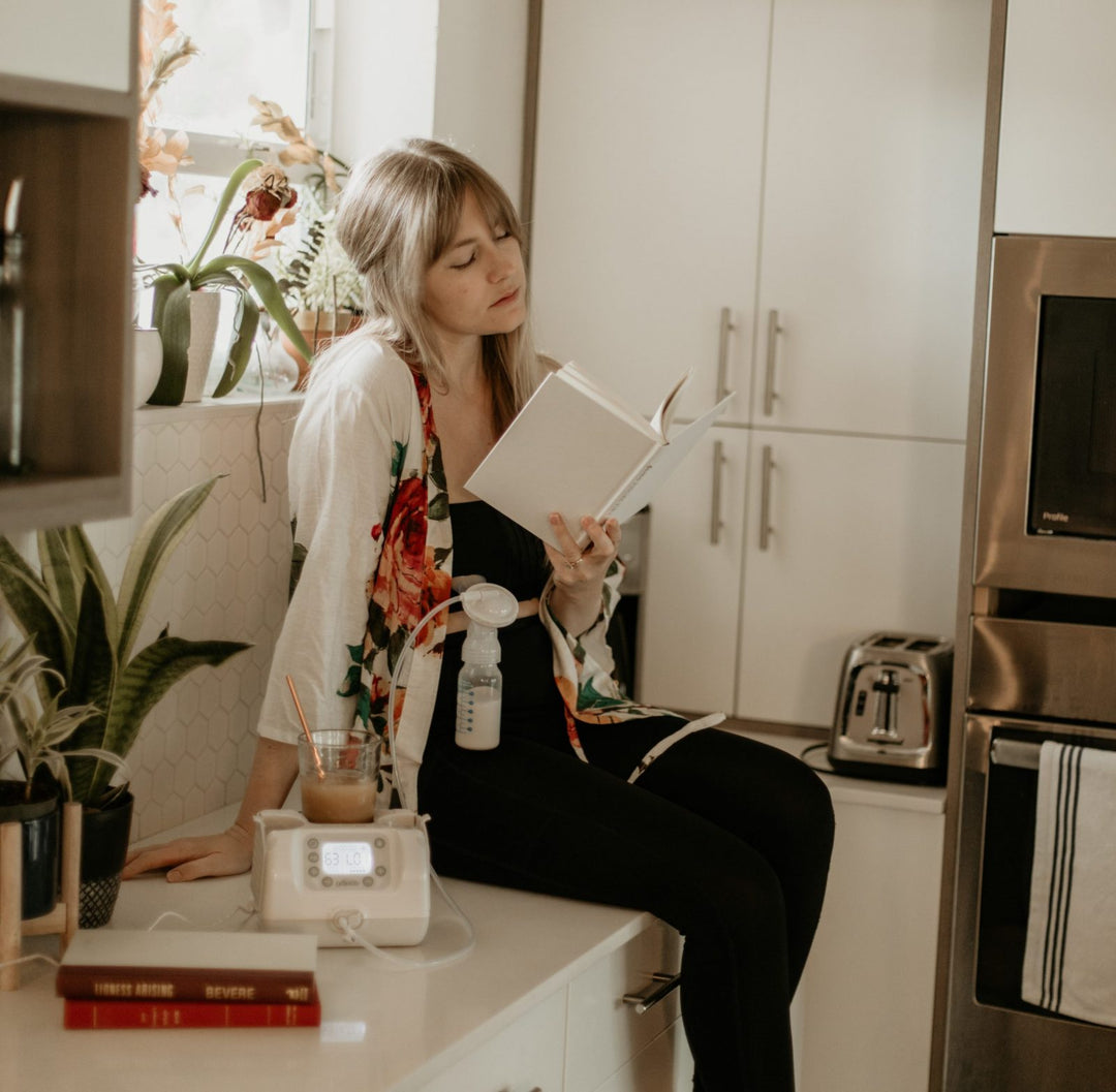 A woman sits on a kitchen counter reading a book, surrounded by potted plants and a breast pump. A glass of juice and a toaster are on the counter beside her. She wears a floral-patterned cardigan and looks focused on her reading.