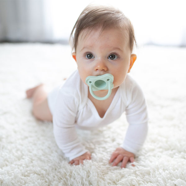 A baby wearing a white onesie is crawling on a fluffy white carpet while looking ahead. The baby has a green pacifier in their mouth. The background is softly blurred, creating a calm and cozy atmosphere.