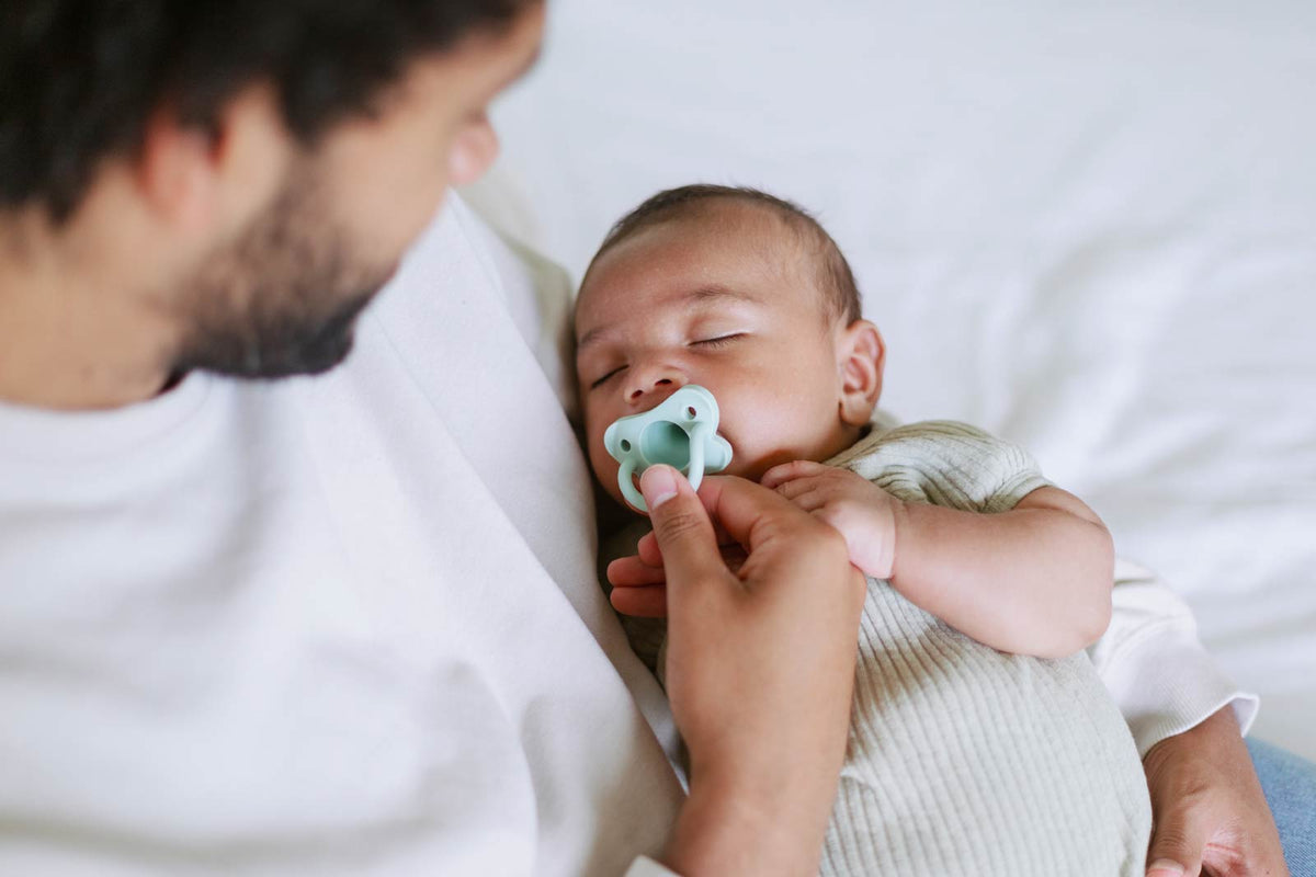 Newborn baby with a Dr. Brown's HappyPaci pacifier being held by an adult in a white setting