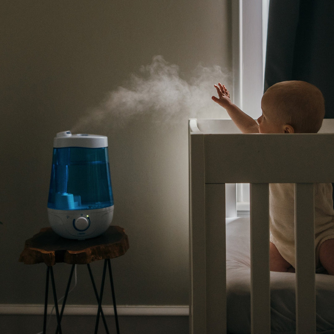 A baby in a crib reaches towards a cool mist humidifier on a small wooden table. Mist fills the air in a softly lit room, creating a serene atmosphere. The crib is white, and the humidifier features a blue water tank.