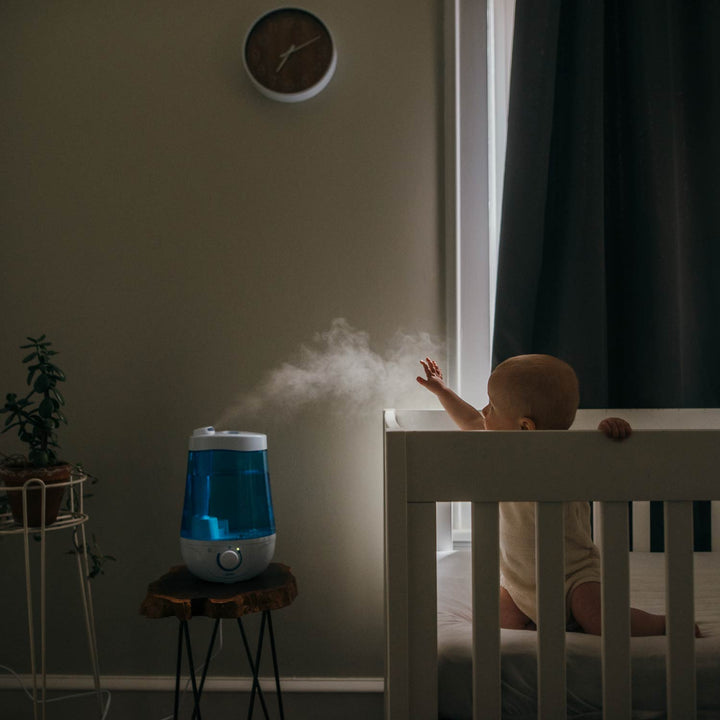 Baby in a crib next to a humidifier with mist in a dimly lit room.