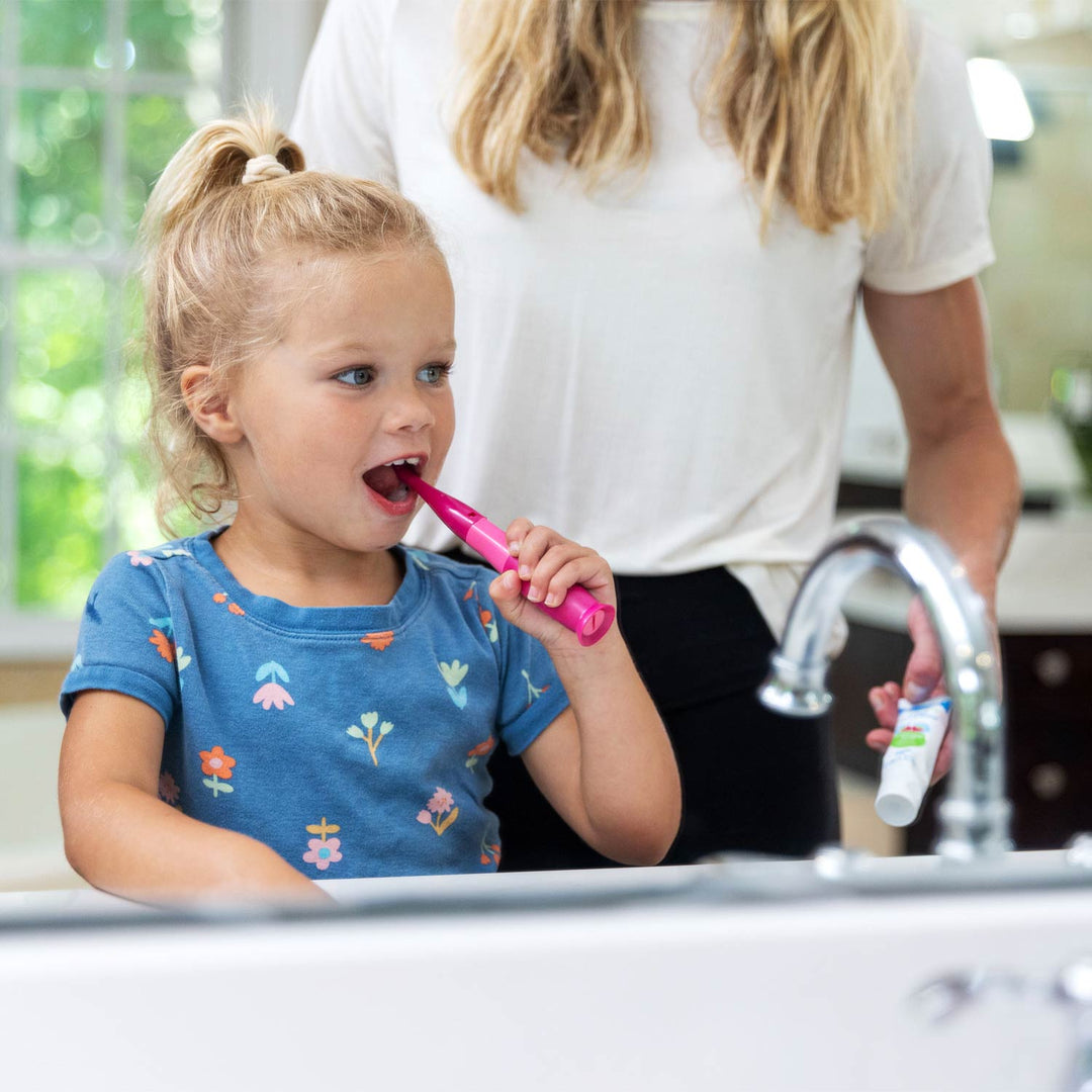 A young blonde girl in a blue floral shirt brushes her teeth with a pink electric toothbrush while standing at a bathroom sink. An adult woman in a white t-shirt stands behind her holding a toothpaste tube.