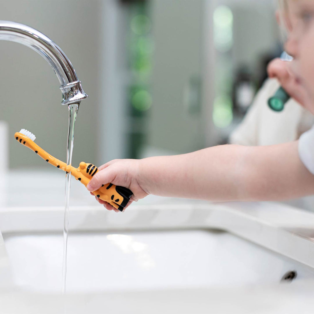 A child's hand holding an orange and black tiger-shaped toothbrush under running water from a chrome faucet in a bathroom sink.