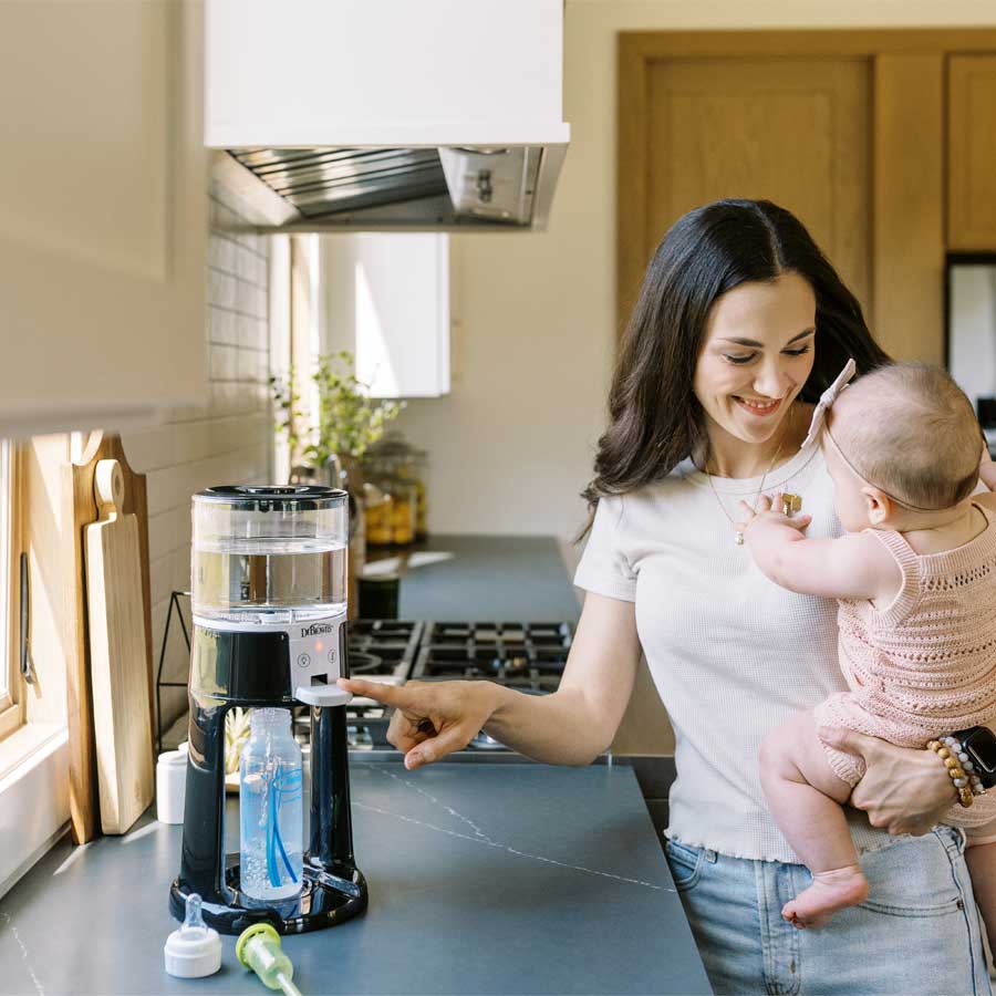 A woman holding a baby in a kitchen uses a baby formula dispenser on the counter. The kitchen is bright and features wooden accents.