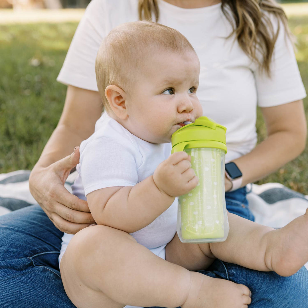 A baby in a white onesie sits on an adult’s lap outdoors, sipping from Dr. Brown’s® Milestones™ Insulated Straw Cup, 2-Pack. They’re on a grassy blanket, enjoying the spill-proof cup; the adult’s face isn’t visible.