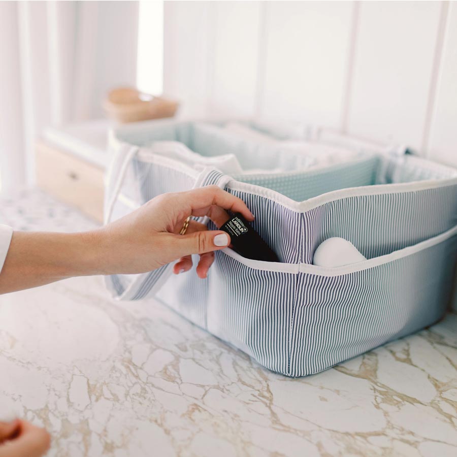 A hand places a small black bottle into a pocket of a striped organizer bag on a marble countertop. The bag has multiple compartments and is part of a light, airy setting with wooden and white decor elements.