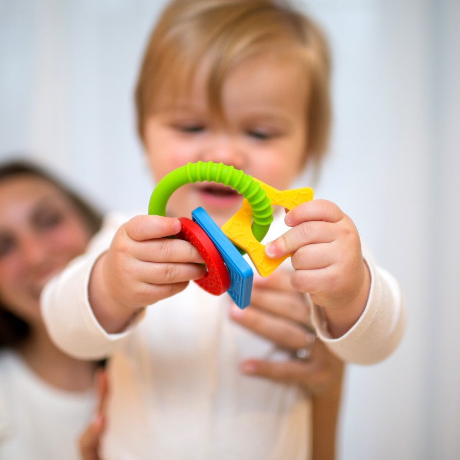 A baby in a white outfit holds a colorful teething ring with different shapes, including a star and circle, while a smiling adult is visible in the background.