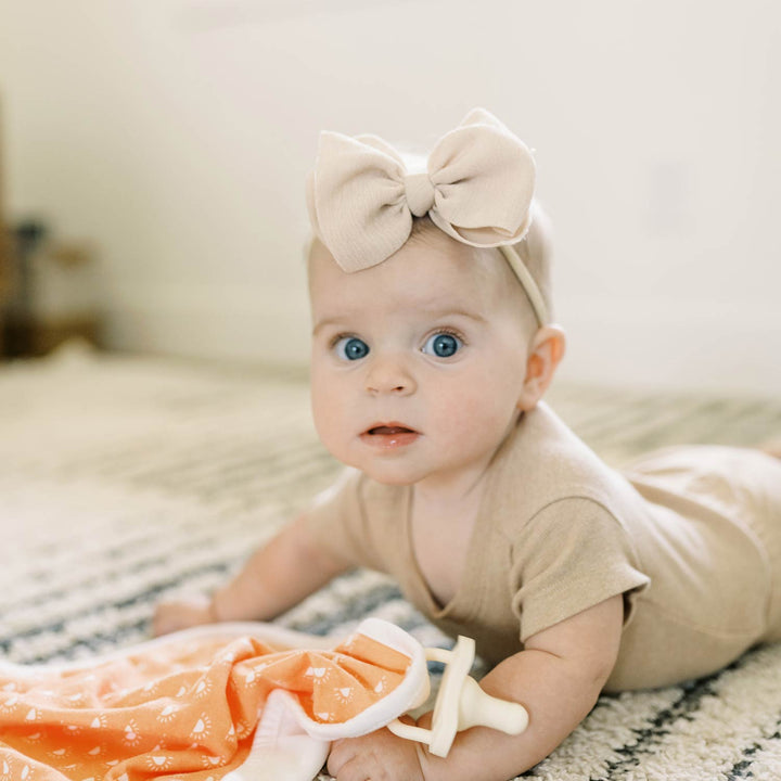 A baby with blue eyes in a beige outfit and bow headband lies on a patterned rug next to a Dr. Brown's™ Lovey Blanket Pacifier and Teether Holder by Dr. Brown's.