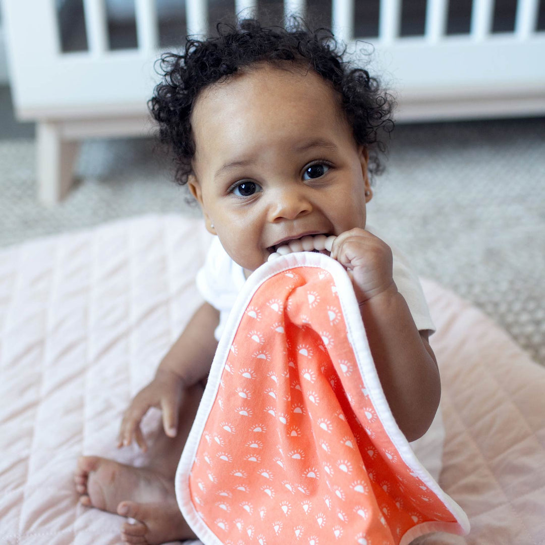 A baby with curly hair sits on a quilted blanket, holding and chewing on Dr. Brown's™ Lovey Blanket Pacifier and Teether Holder. A white crib is visible in the background.