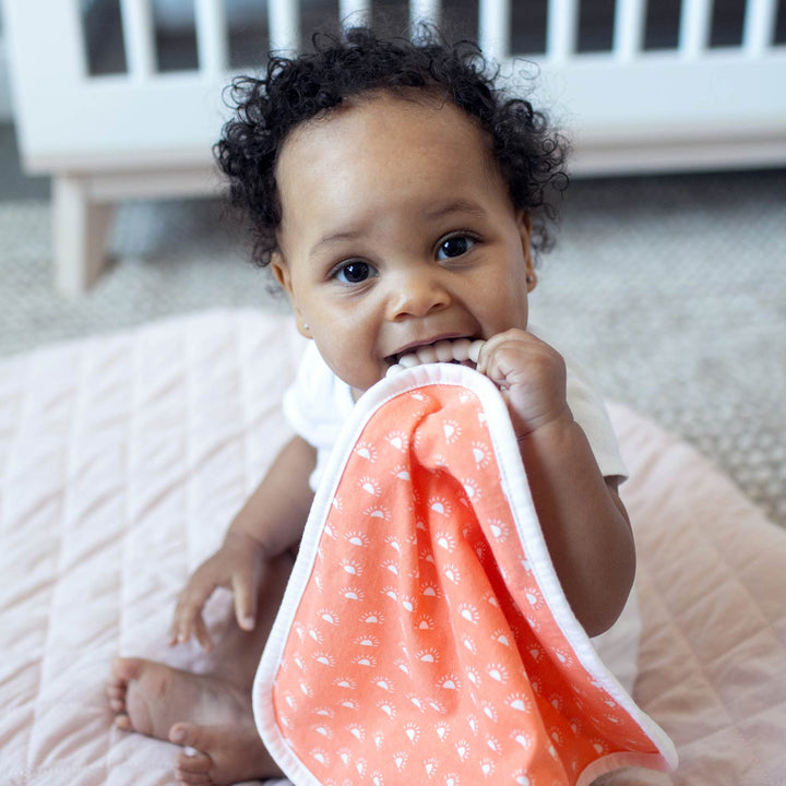 A baby with curly hair sits on a quilted blanket, holding and chewing on Dr. Brown's™ Lovey Blanket Pacifier and Teether Holder. A white crib is visible in the background.