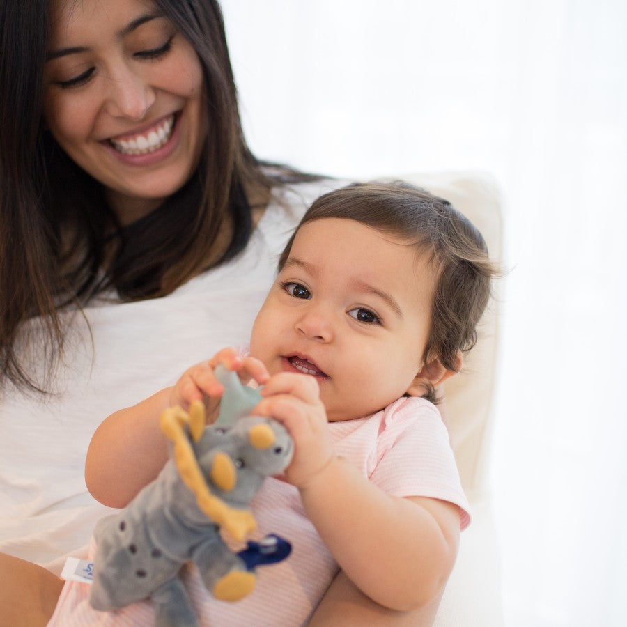 A woman smiles while holding a baby in her lap. The baby, wearing a light pink outfit, looks at the camera and holds a gray stuffed elephant toy. The background is softly lit and blurred.