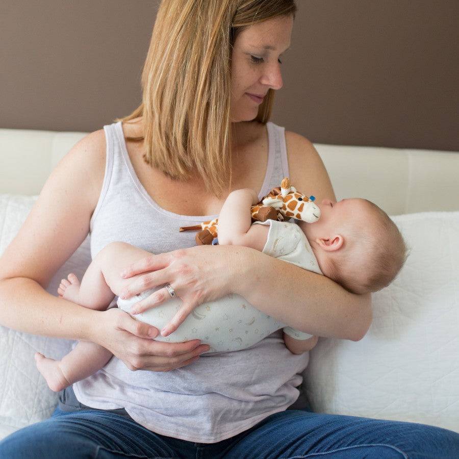 A woman is sitting on a bed, smiling down at a baby she is cradling in her arms. The baby, wearing a white onesie, holds a plush giraffe toy. The background is a neutral-colored wall and a white pillow.