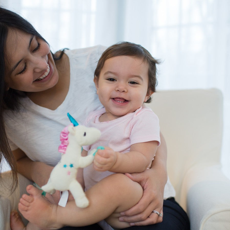 A woman smiles at a baby sitting on her lap, holding a small stuffed unicorn. The scene is in a bright room with natural light coming through the windows. The baby is wearing a pink shirt and looks content.