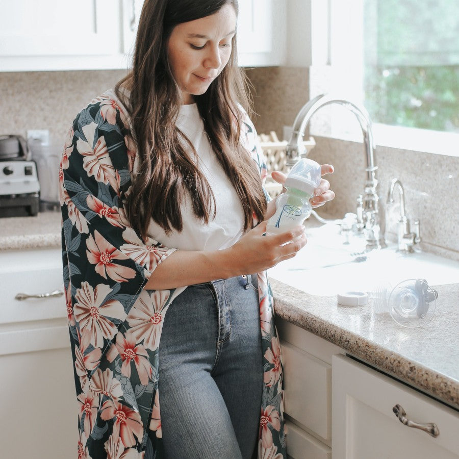 A woman in a floral kimono stands by a kitchen counter, holding a baby bottle with a green lid. She is looking down at the bottle. The kitchen has a sink, toaster, and various items on the counter. Sunlight shines through a window.