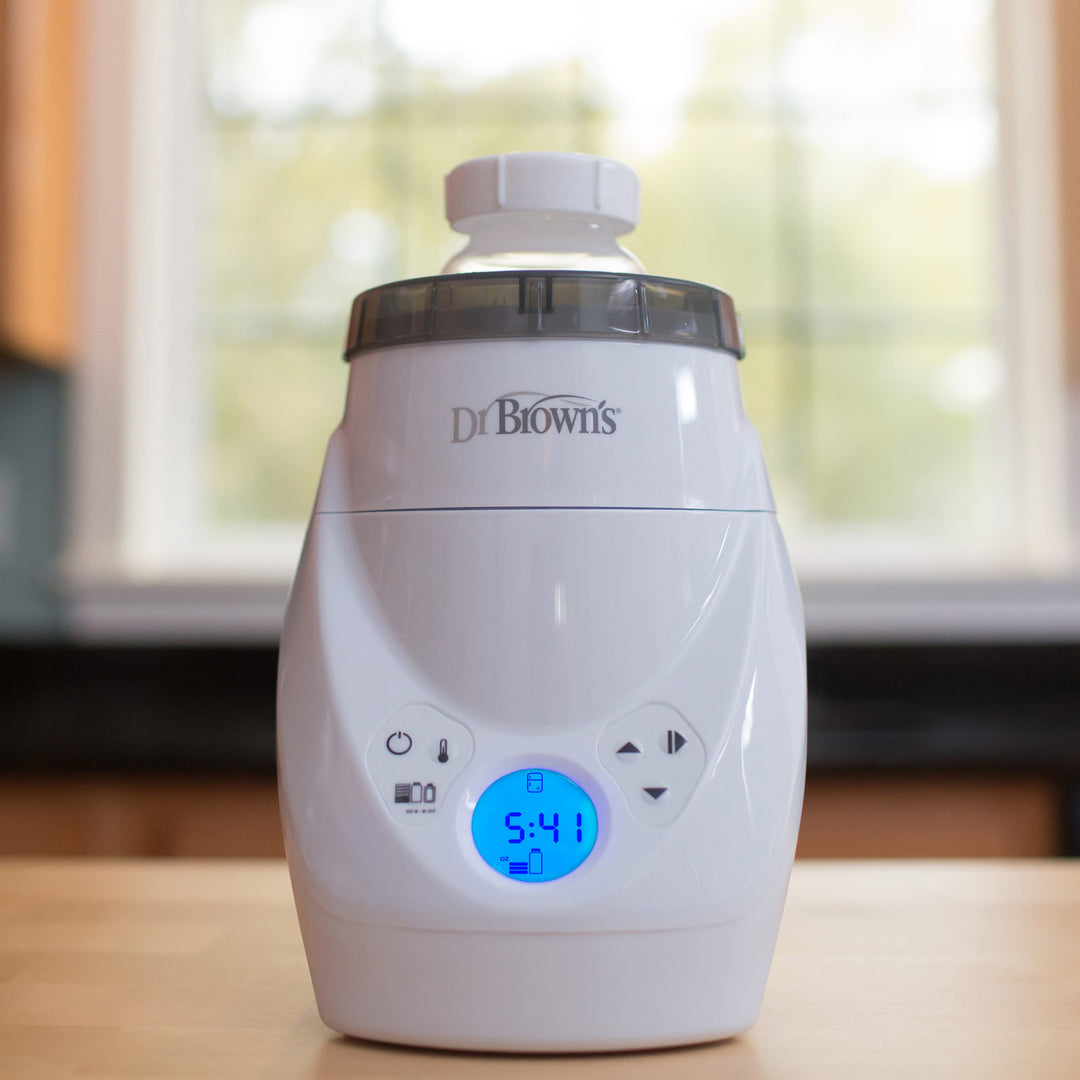 A Dr. Brown's bottle warmer is placed on a wooden surface with a bottle on top. The digital display on the front shows the time 5:41. The background reveals a softly blurred window view.