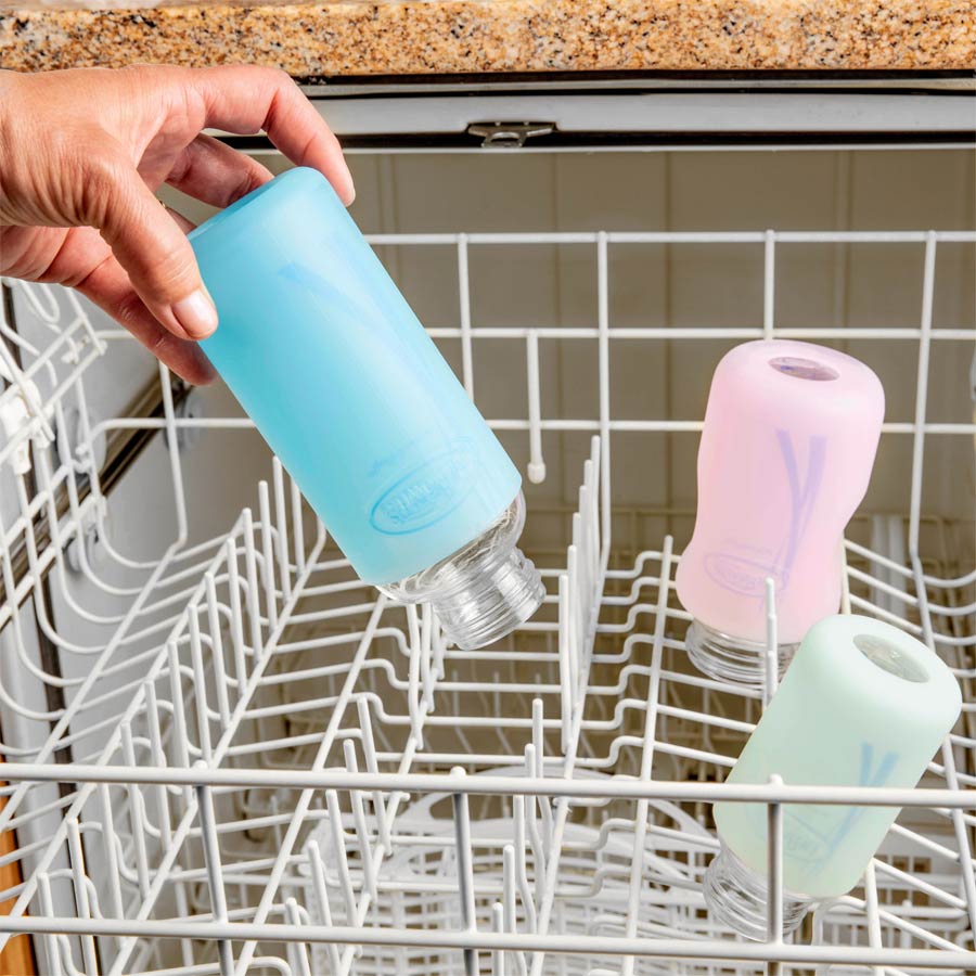 A person places a blue silicone-covered glass bottle upside down in a dishwasher rack. Two other similar bottles, one pink and one mint green, are already positioned in the dishwasher. The countertop above is made of brown speckled material.