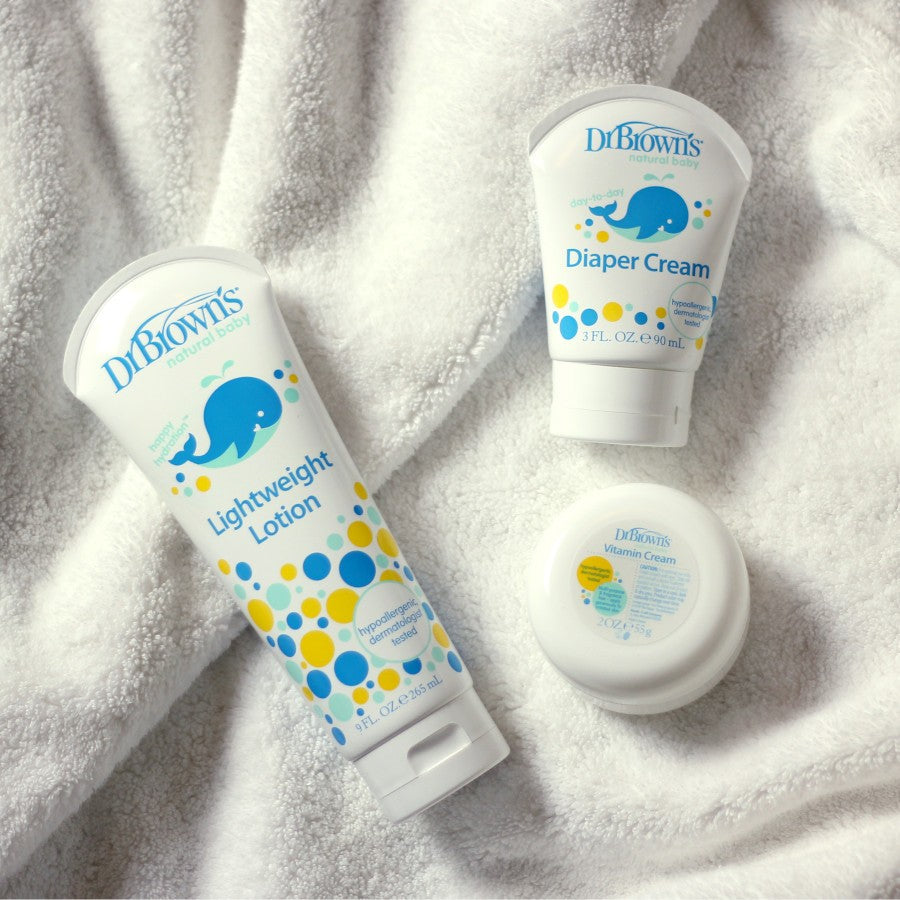 Three Dr. Brown's baby care products on a soft white towel: Lightweight Lotion, Diaper Cream, and a container of Vitamin Cream. Each product features playful blue and yellow designs, with the lotion tube and cream bottle displaying a blue elephant.