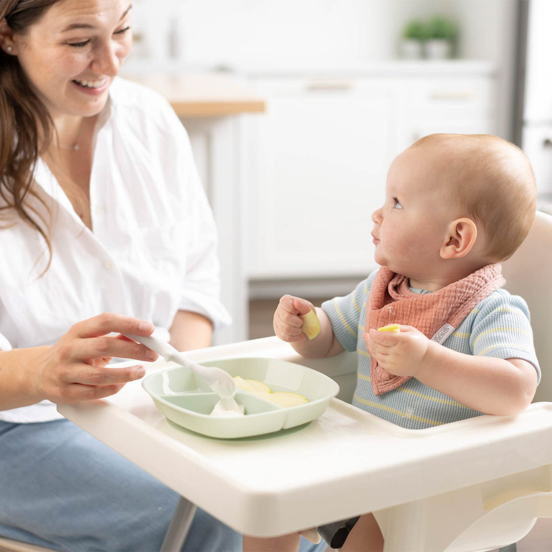 A baby sitting in a high chair wearing a striped shirt and a pink bib, eating from a light green divided plate with a white spoon held by a smiling woman in a white blouse.