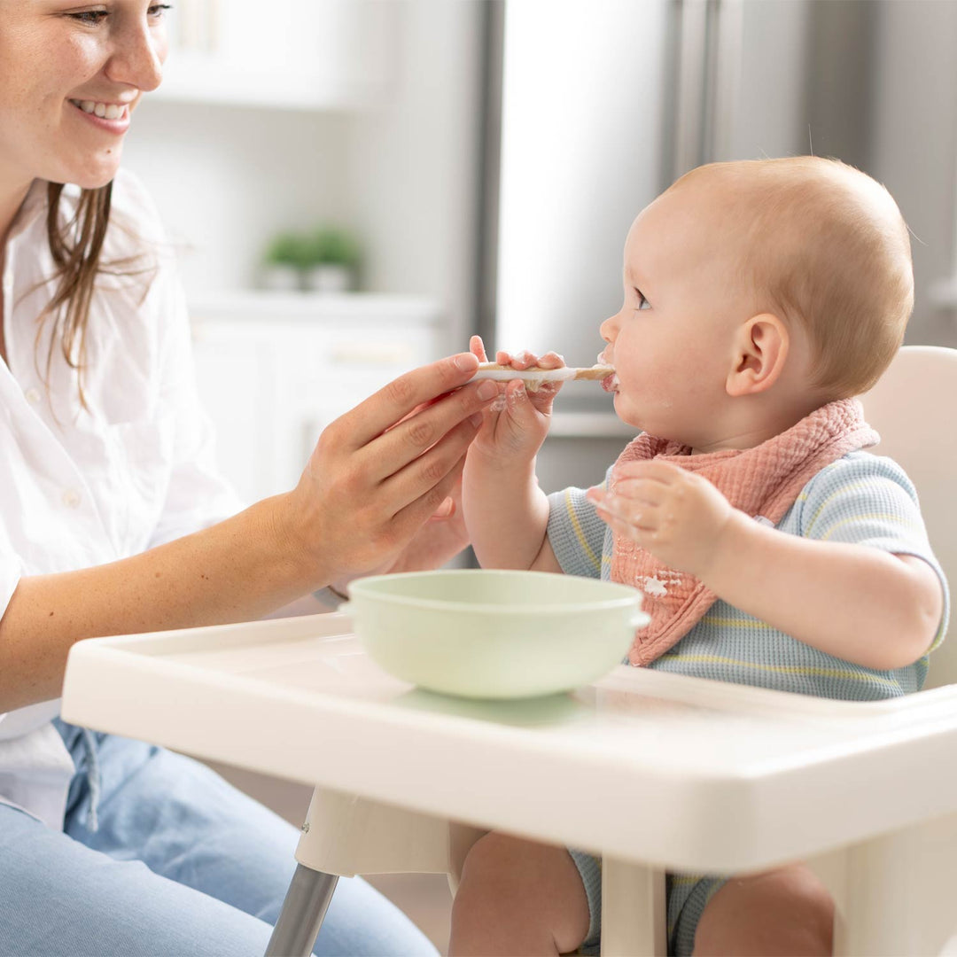 A baby in a striped blue and white outfit with a pink bib sits in a white high chair while a woman feeds the baby with a spoon. A light green bowl is on the chair tray in a bright kitchen setting.