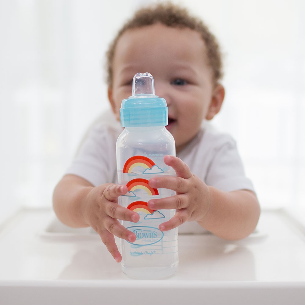 A baby with curly hair is sitting at a high chair, holding a Dr. Brown's baby bottle decorated with rainbows and clouds. The baby is wearing a white shirt and reaching for the camera with a happy expression.