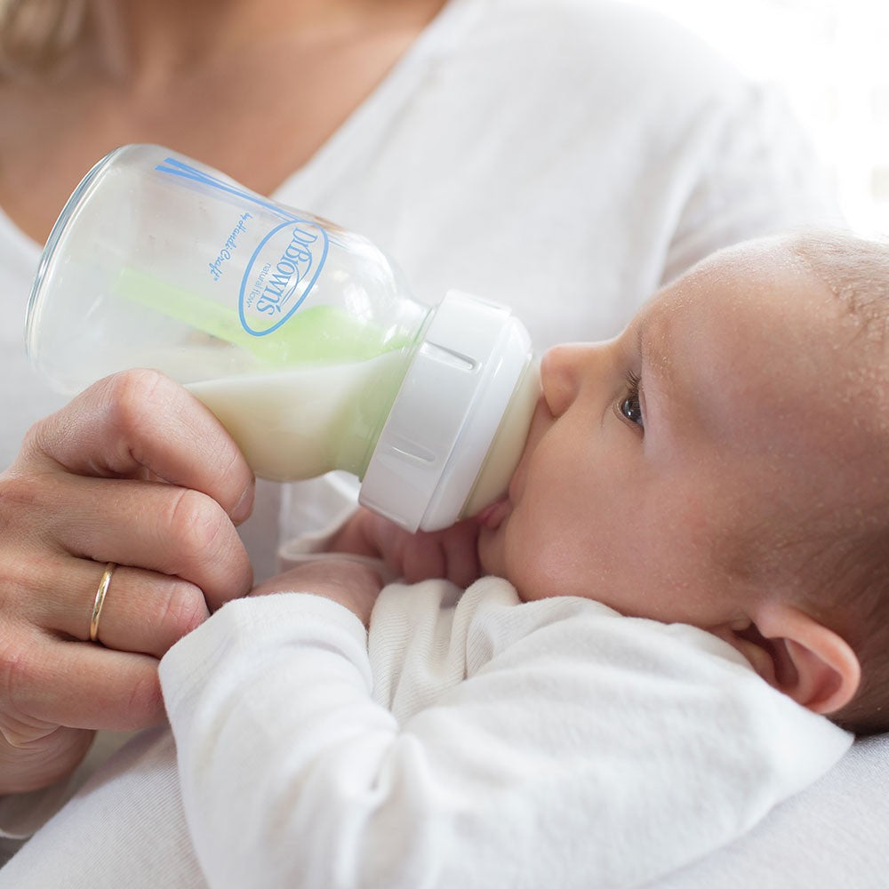 A baby drinking milk from a bottle while being held by an adult. The baby is wearing a white outfit, and the adult's hand is gently supporting the bottle. The scene conveys a calm and nurturing moment.